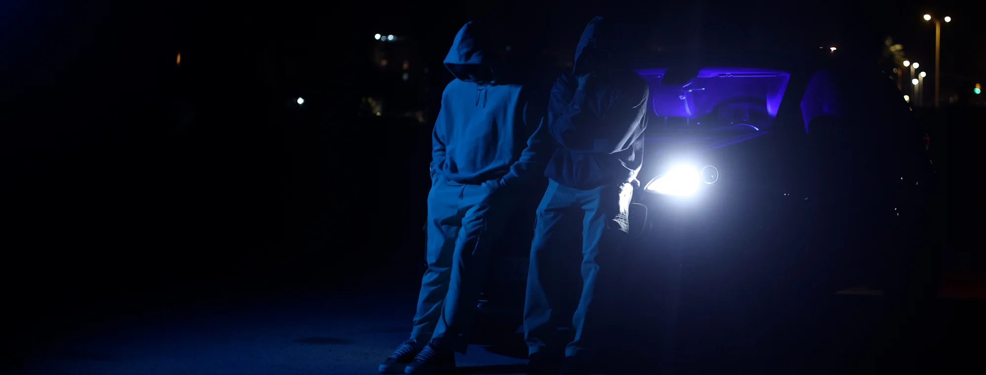 Dos artistas de noche, apoyados en un coche con los faros encendidos